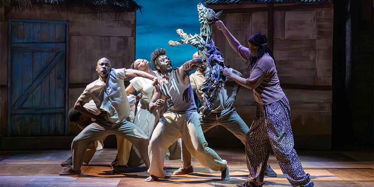 Five performers engage dynamically on stage, with one holding a puppet amidst a textured backdrop.