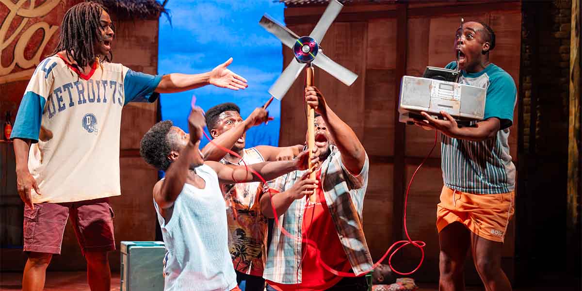 Five actors engage energetically, one holding a fan, while another operates a device, expressing excitement.