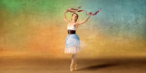 A dancer in a light blue dress performs against a multicoloured background holding a red ribbon above their head.