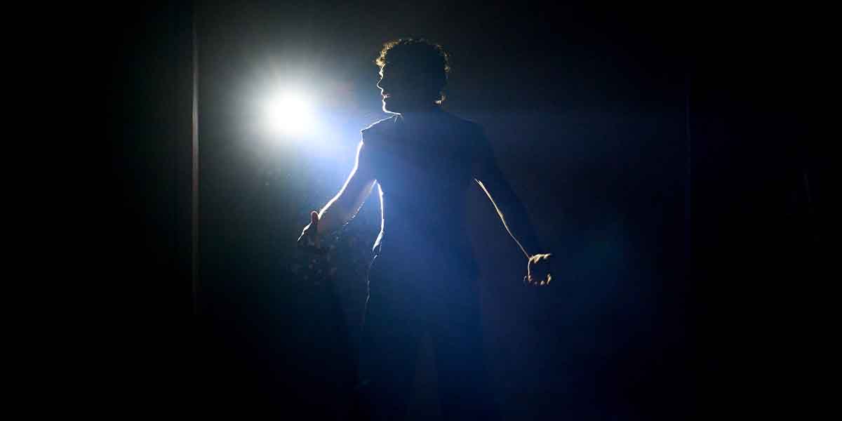 Noah Jupe stands in silhouette, arms raised, illuminated by a bright spotlight against a dark background.