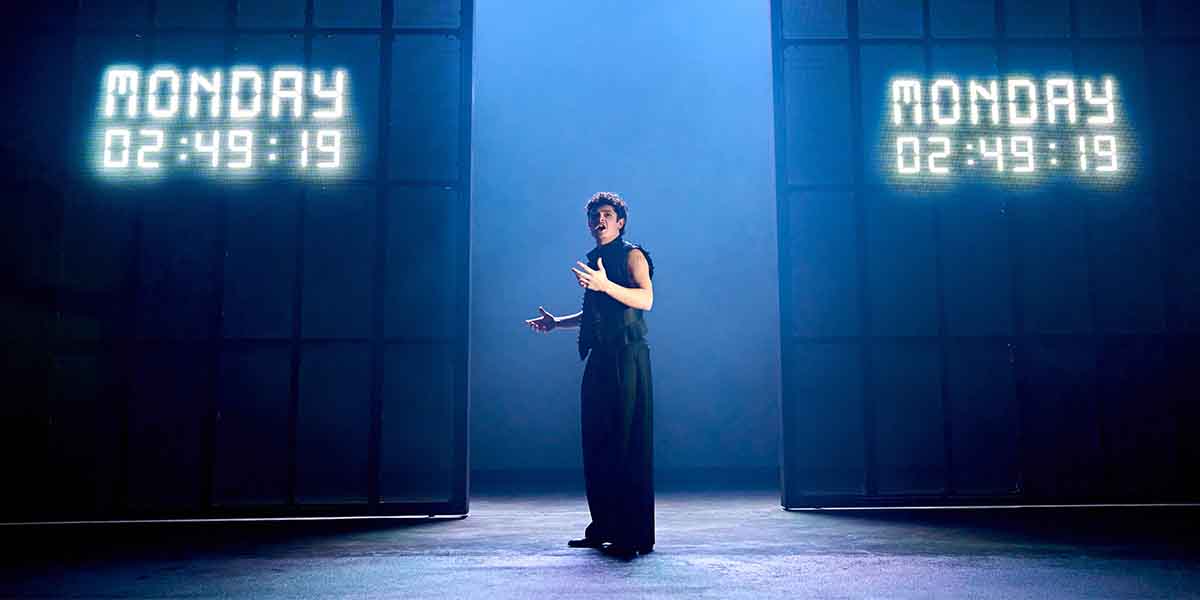 Noah Jupe stands centre stage, gesturing, with illuminated digital clocks displaying "MONDAY 02:49:19" in the background.