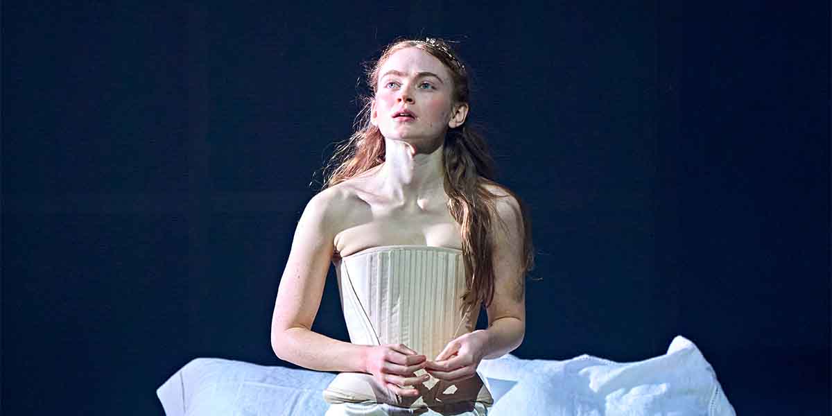 Sadie Sink, dressed in a light corset, gazes upwards against a dark background, conveying emotion.