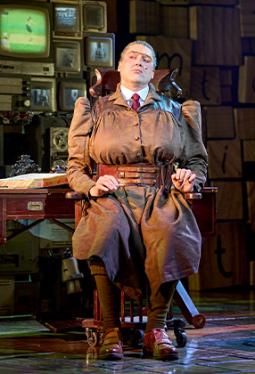 A character in a large, brown costume sits at a desk, surrounded by vintage televisions and boxes.