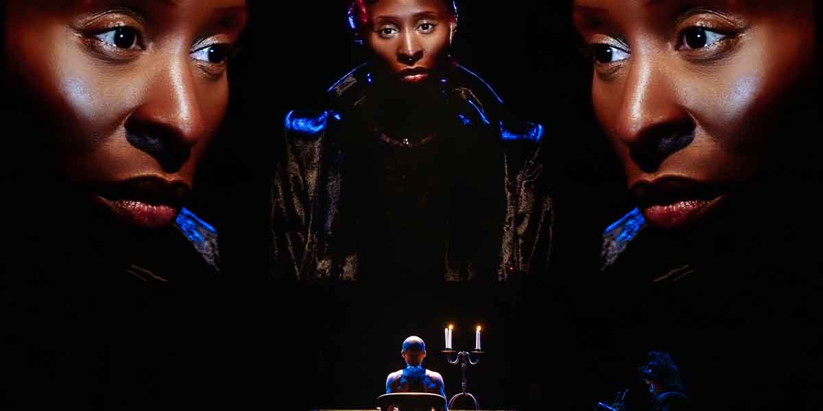 A woman with dark skin and striking features sits at a table, illuminated by candlelight, with large projections of her face behind.
