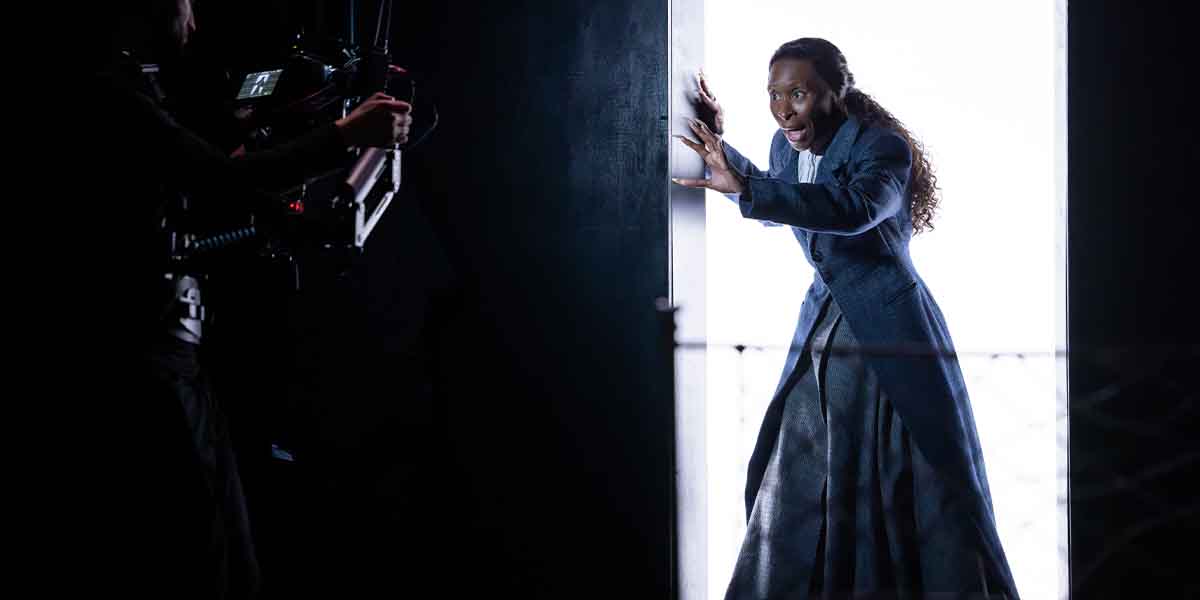 A person in a dark costume expresses surprise while standing in a doorway, illuminated from behind.