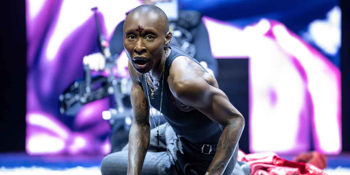 A performer in a black tank top kneels on stage, looking intensely towards the audience, with dramatic lighting.