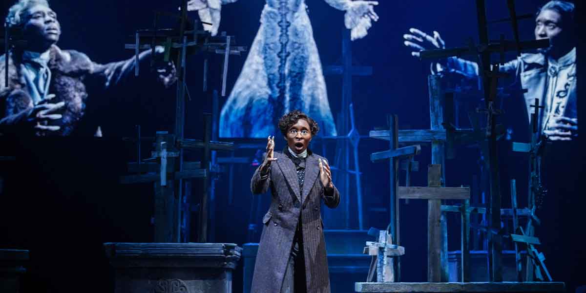 A performer in a dark coat gestures dramatically, with ghostly figures and wooden crosses in the background.