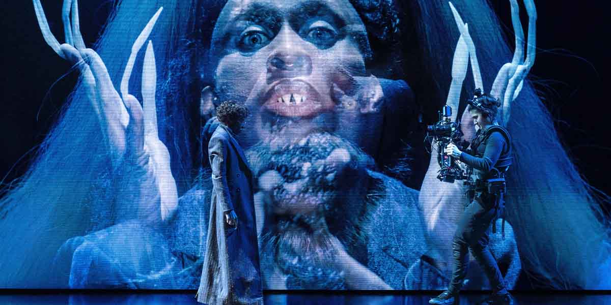 A performer in a long coat stands before a large, eerie projection of a face with fangs.