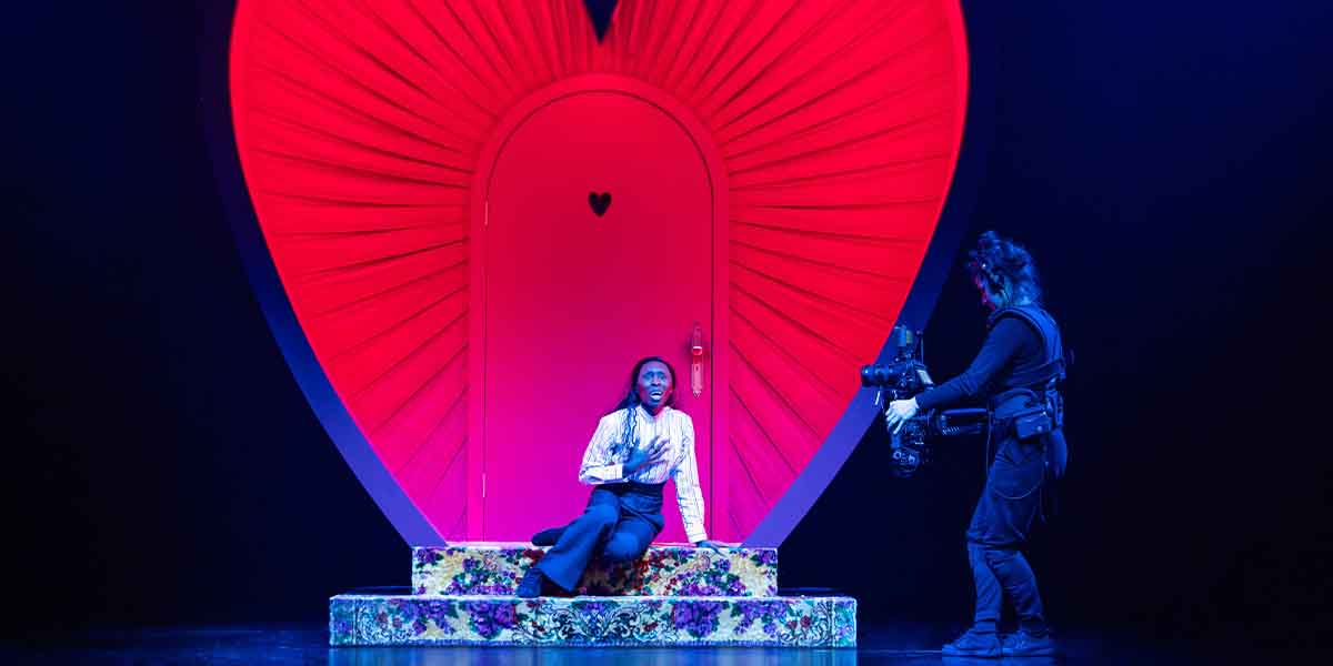 A performer in a white shirt sits on floral steps, expressing emotion, while another person holds a camera.