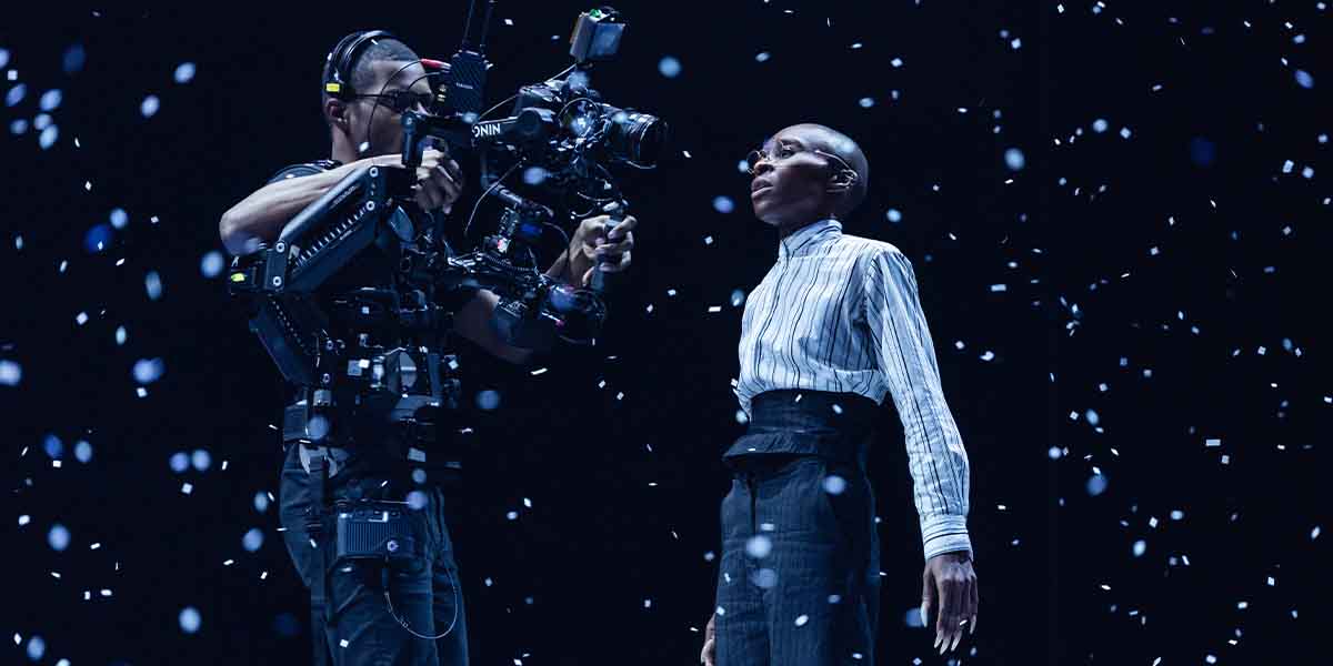 A camera operator films a performer in a striped shirt, surrounded by falling snowflakes on stage.