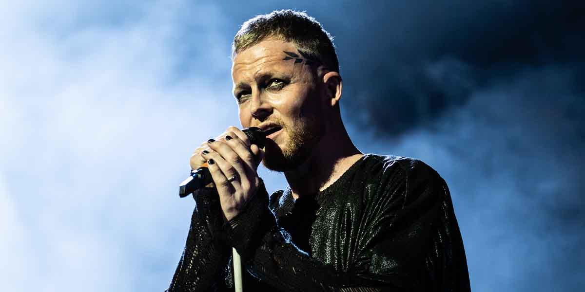 David Thaxton performs passionately, holding a microphone, wearing a textured black outfit against a smoky background.