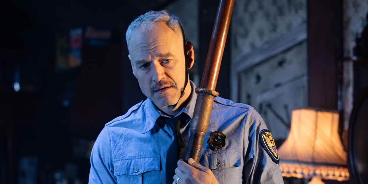 A person in a blue police uniform holds a wooden baton, looking serious in a dimly lit room.