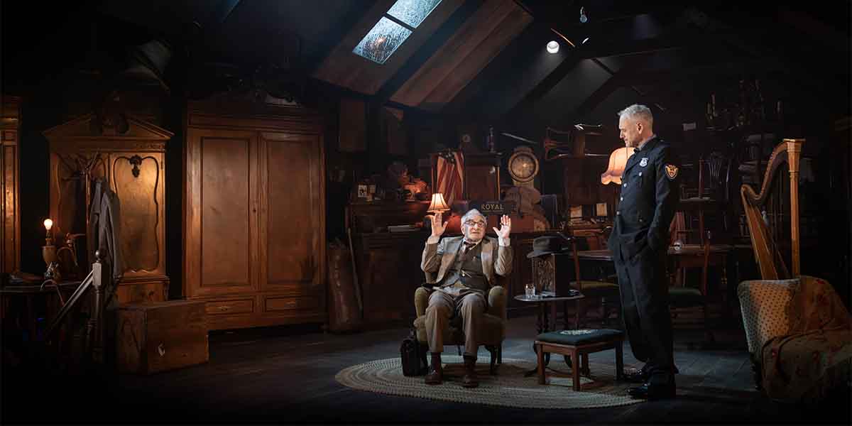 A person in a suit gestures expressively while seated, as a police officer stands nearby in a dimly lit room.