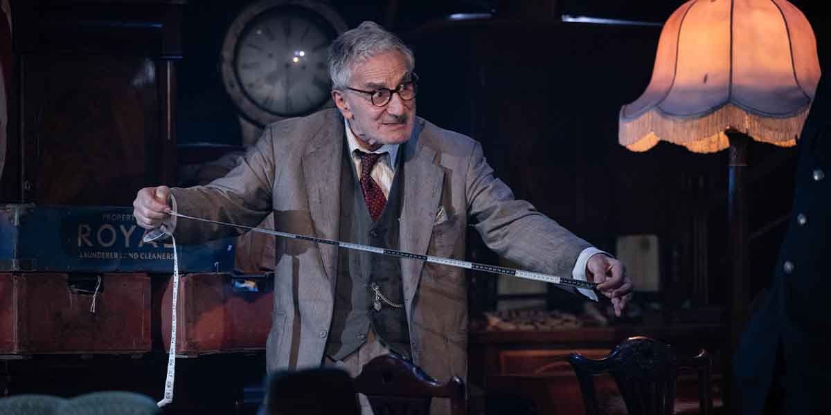 A person in a grey suit holds a measuring tape, standing in a dimly lit room with vintage decor.