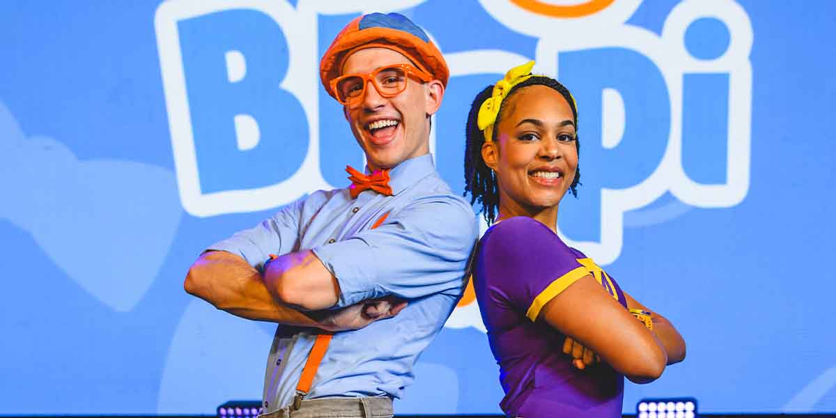 Two performers pose back-to-back, smiling. One wears orange glasses and a blue shirt, the other a purple outfit.