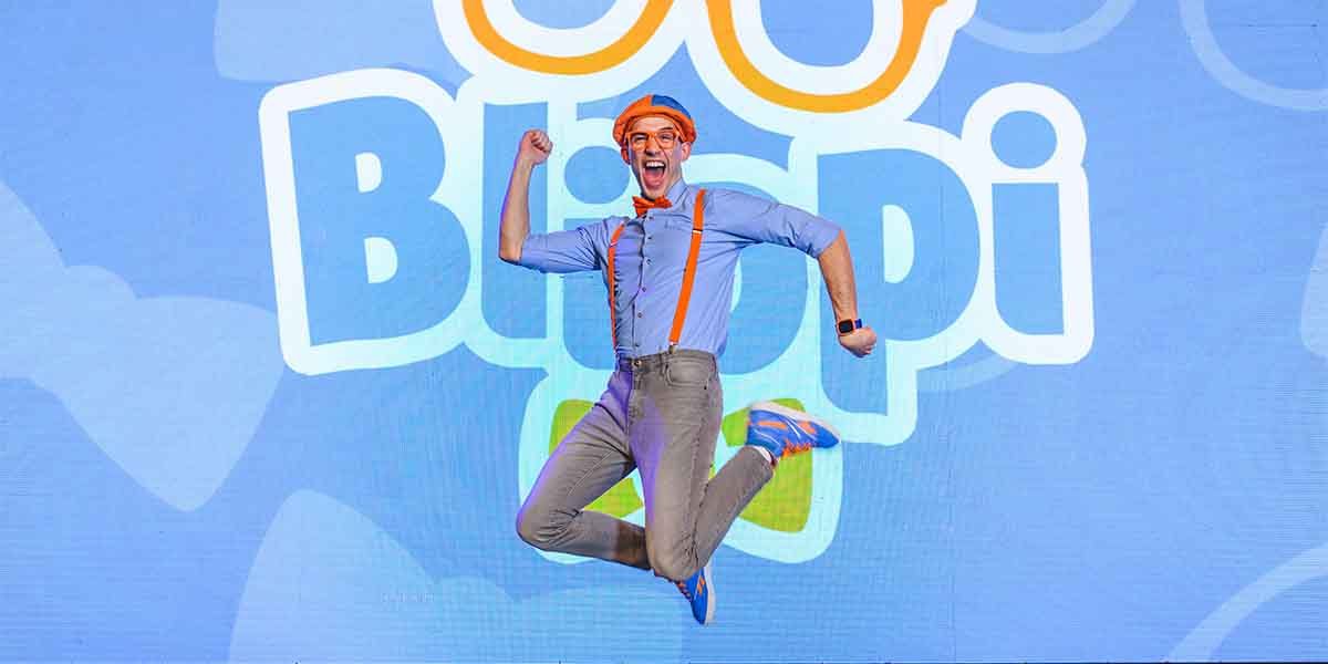 A man in a blue shirt and orange accessories jumps joyfully against a colourful "Be Like Blippi" backdrop.