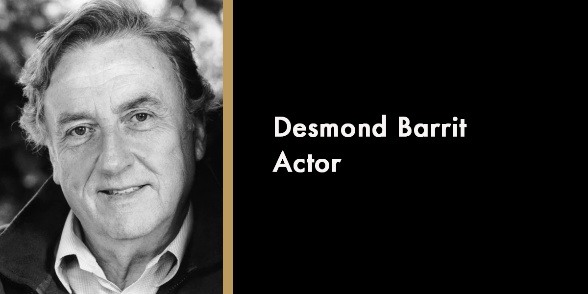 A portrait of Desmond Barrit, smiling, with a dark background and soft lighting highlighting his features.