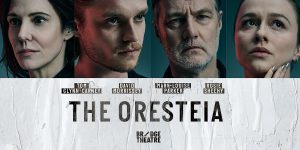 Two ticket agents stand behind a counter, displaying promotional artwork for "The Oresteia" in vibrant colours.