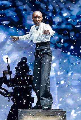 A performer in a white shirt and black trousers stands on a box, surrounded by falling snowflakes.
