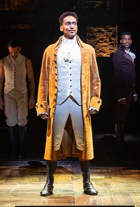 A performer in a brown coat and white attire stands confidently, with two others in the background.