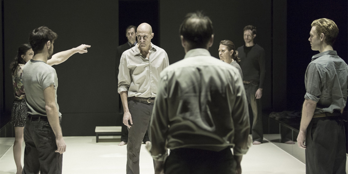 Mark Strong and cast members stand in dramatic poses on stage, with a minimalist black background.