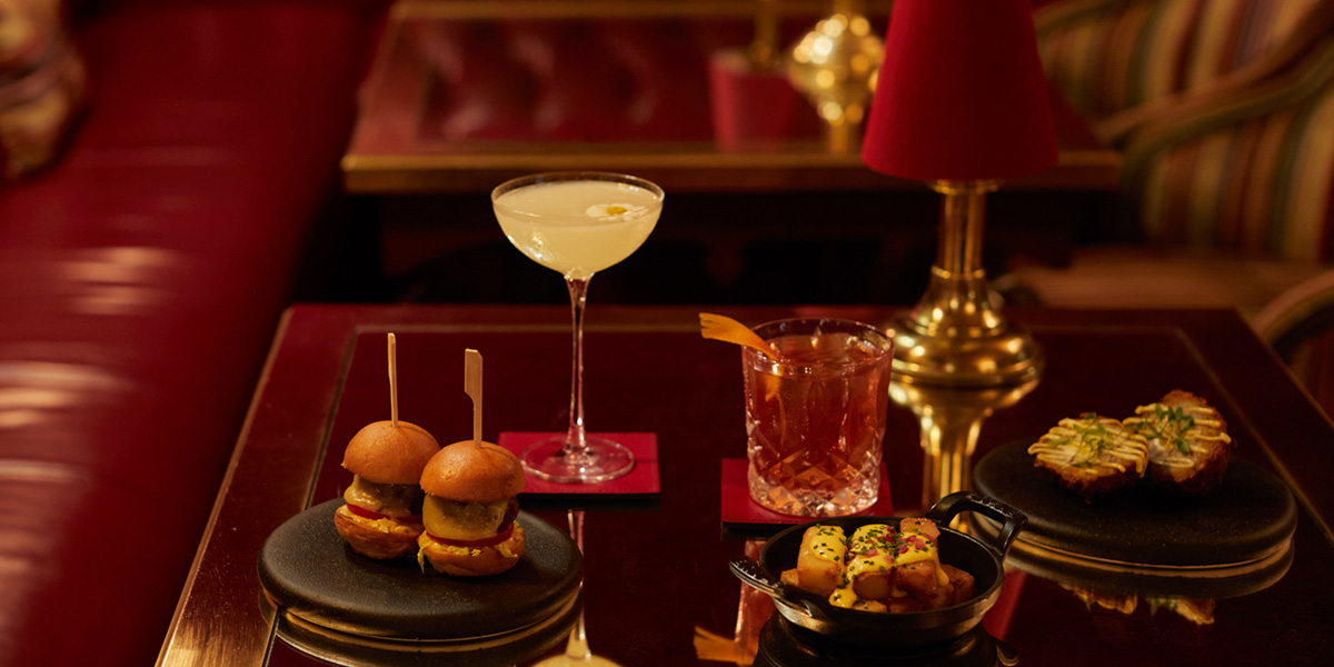 A table displays two sliders, a cocktail, a whiskey drink, and a plate of savoury snacks.