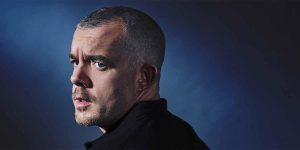A person with short, grey hair gazes over their shoulder, set against a dark blue background.